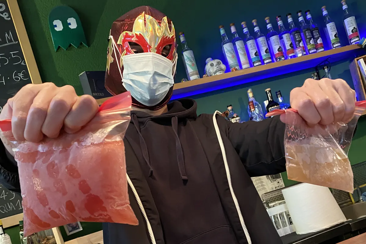 In a luchador mask behind the bar, holding up two bags of frozen cocktails